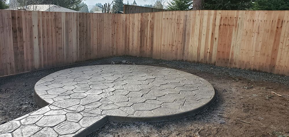 A circular, stone patio with a walkway, set in a backyard against a wooden fence.