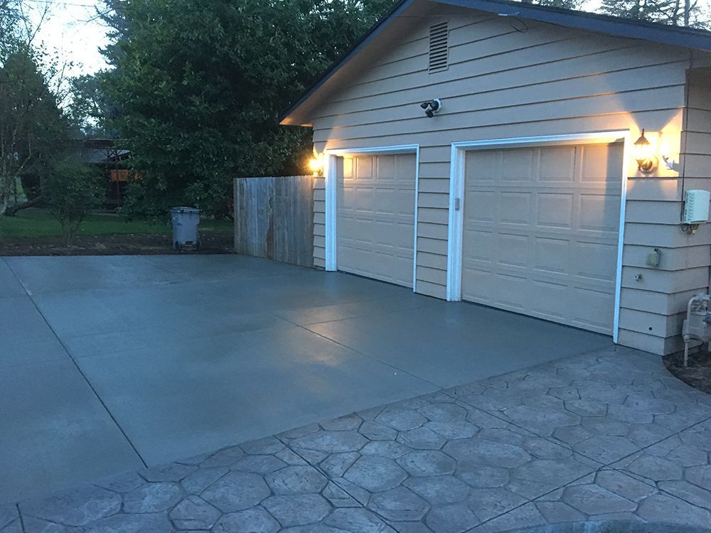 Gray concrete driveway leading to a tan garage with two doors, lit by exterior lights.