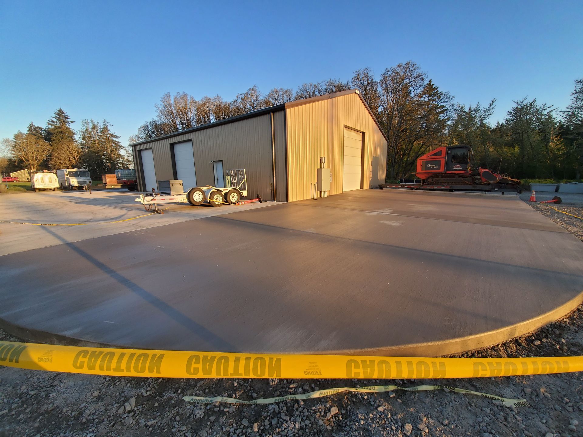 New concrete pad in front of a metal building. Caution tape across the foreground. Construction equipment visible. Blue sky.