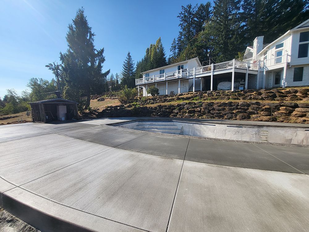 Newly poured concrete patio and pool area next to a large white house on a hill.