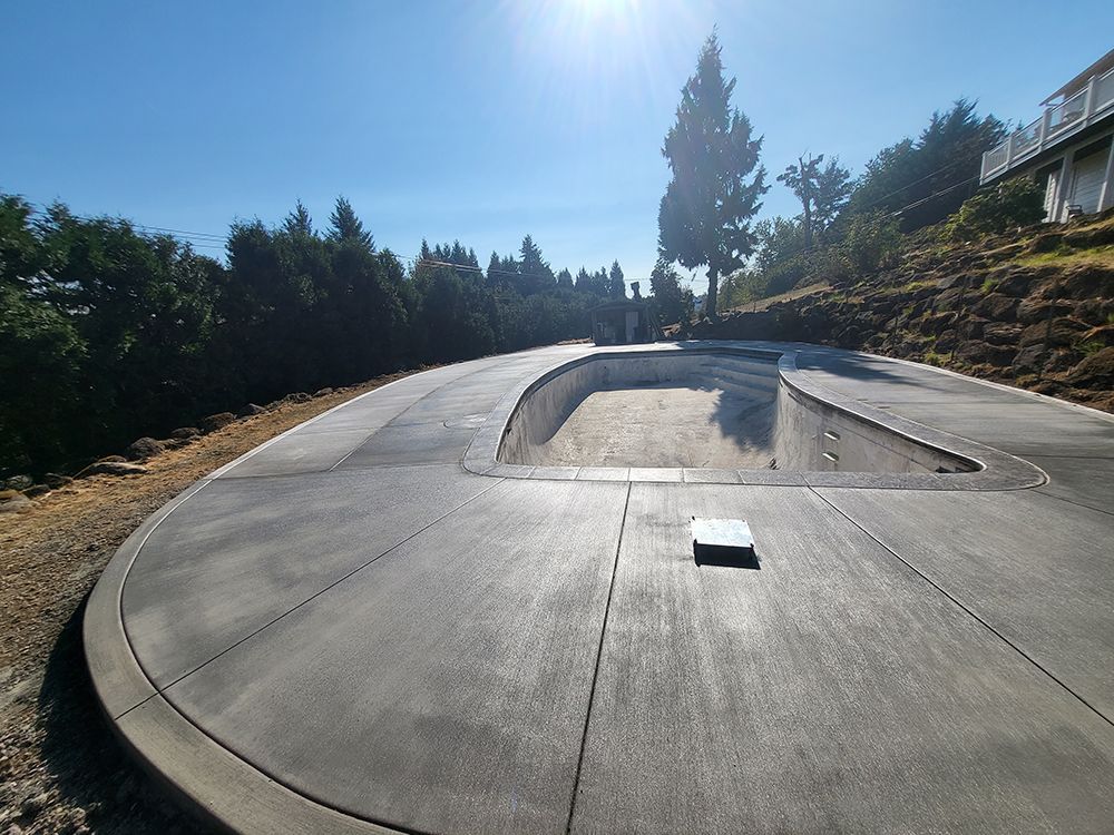 Concrete pool deck with curved edges, under a bright sun. Trees and a house are visible in the background.