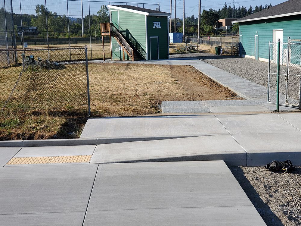 Concrete sidewalk with ramp leading towards a green building, chain link fence and a baseball field.