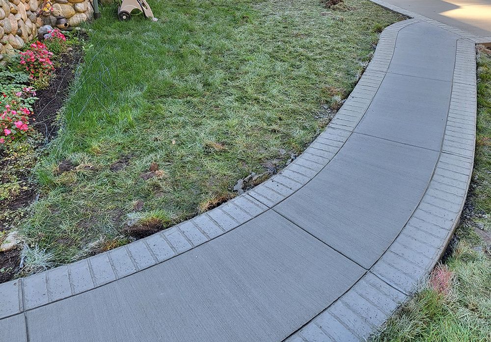 Curved concrete sidewalk bordering a lawn and flowerbed. Gray concrete with decorative edge.