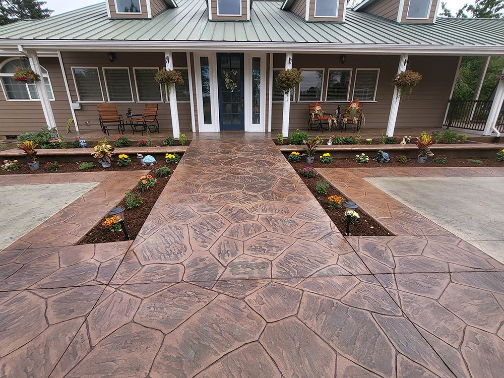 Stamped concrete walkway leading to a house with brown siding, flower beds, and a covered porch.