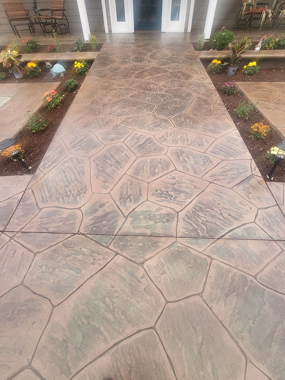 Stamped concrete walkway leading to a doorway, flanked by flowerbeds with colorful flowers.