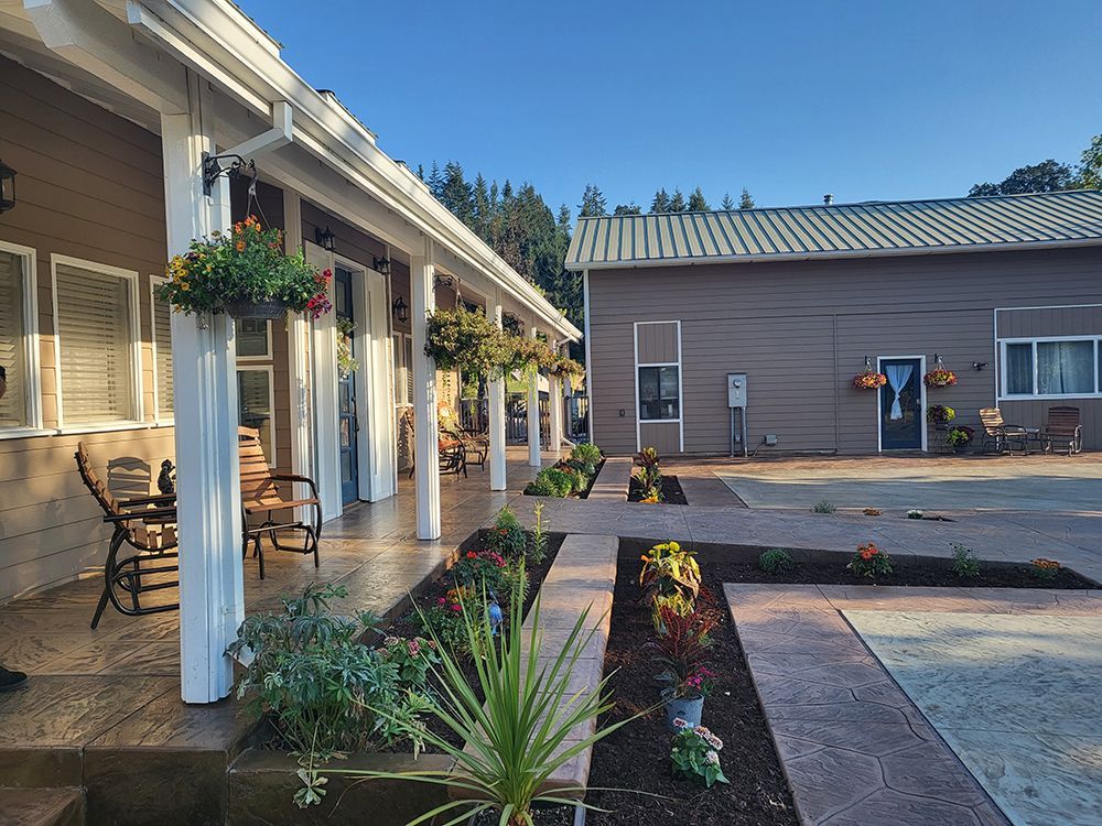 A building with a porch lined with flowers and chairs, with a second building behind it.