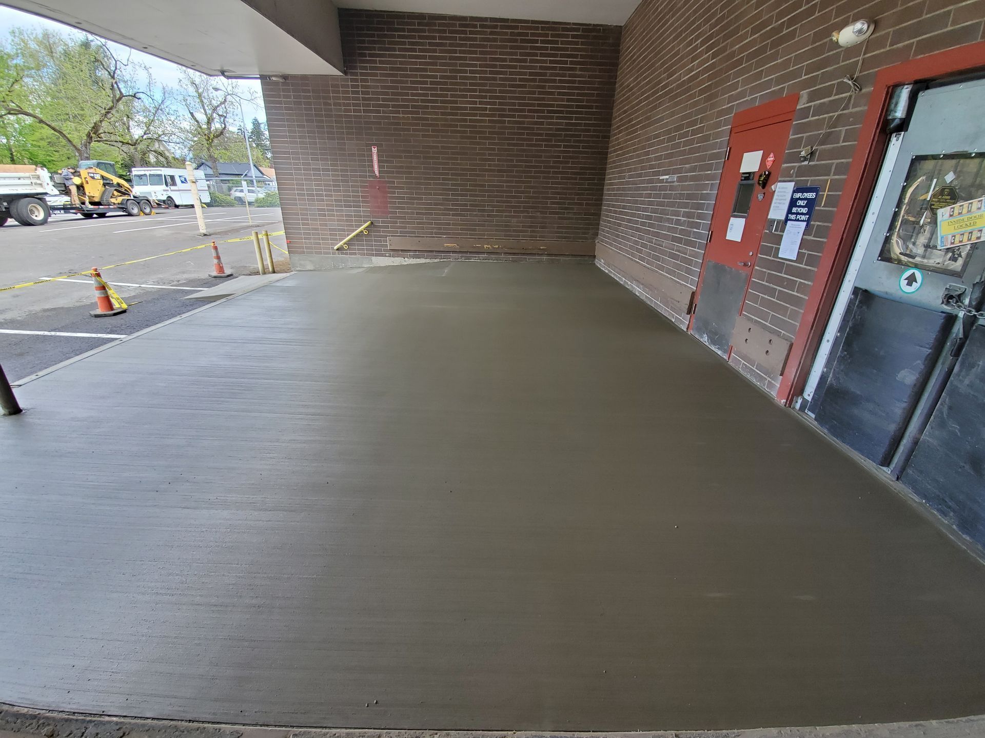 Newly poured concrete surface outside a brick building with two doors. Construction vehicles visible in the background.
