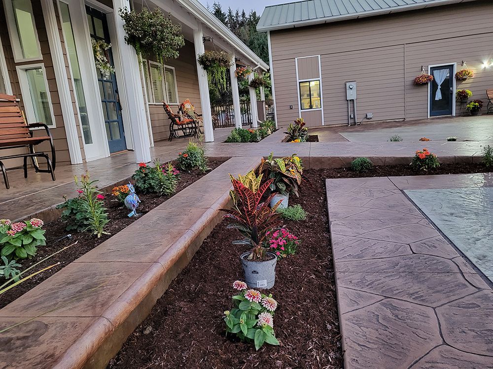 Front yard with concrete pathway, flowerbeds, and a house with a porch and hanging plants.