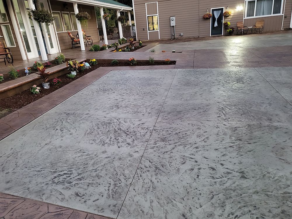 Gray stamped concrete driveway in front of a building with a porch, surrounded by plants and a garden bed.