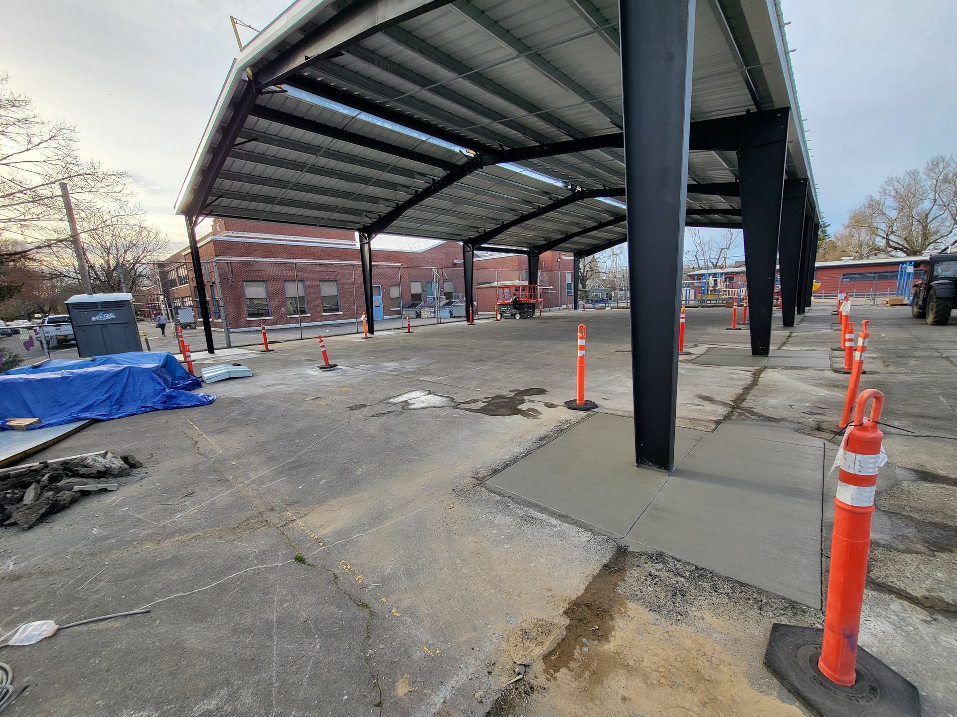 Metal canopy over concrete, asphalt area. Construction site with orange safety cones, brick building in the background.