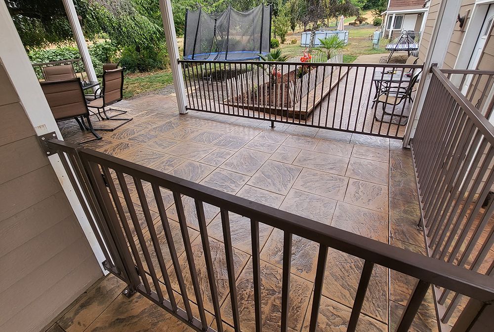 Brown metal railing surrounds a concrete patio with outdoor furniture, overlooking a backyard with a trampoline.