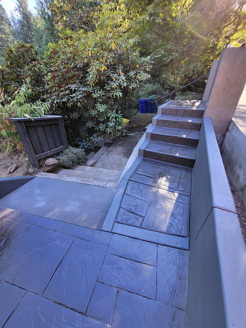 Steps and pathway leading up a hillside, with dark gray stonework, trees, and blue trash can.