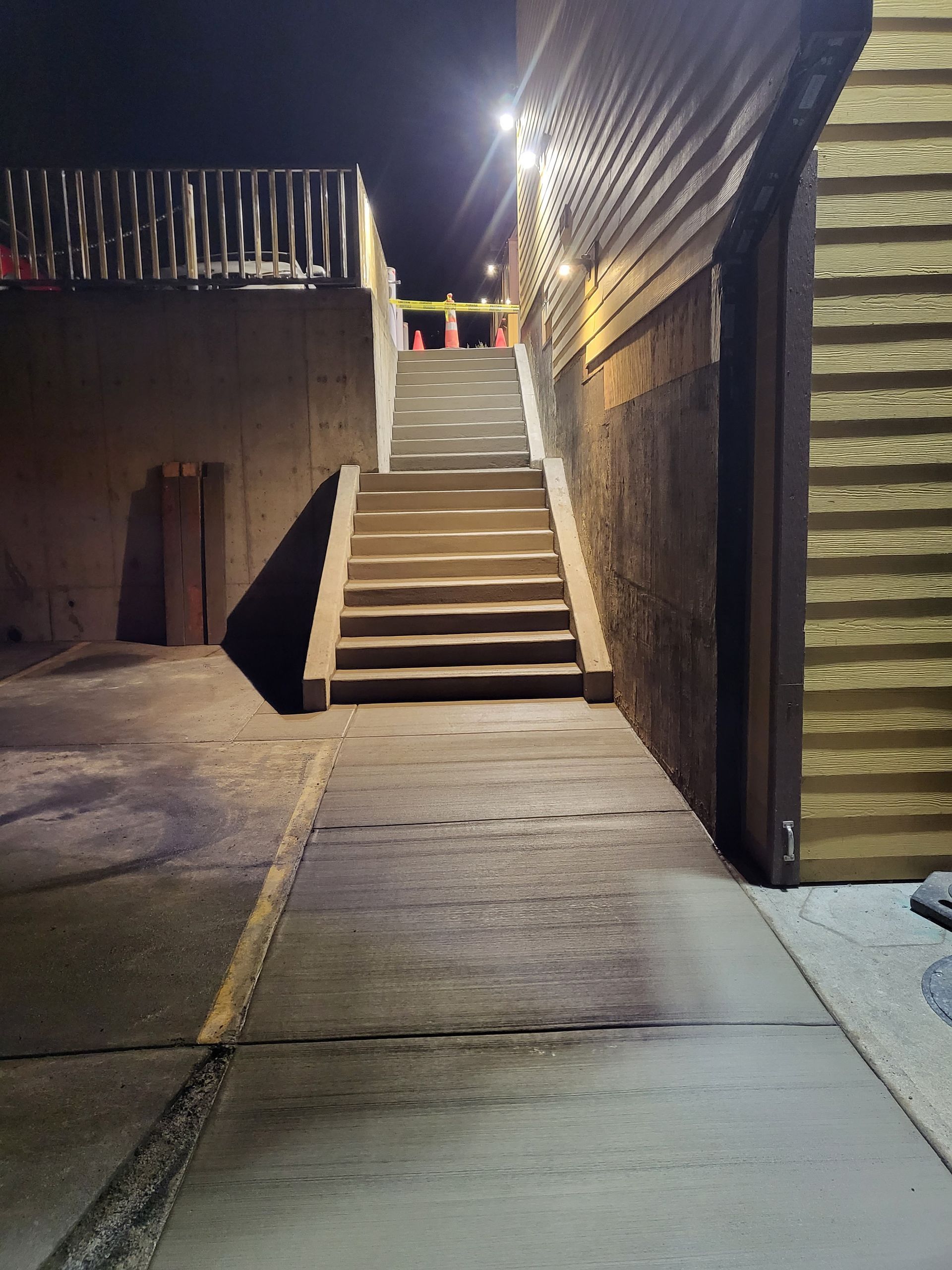 Concrete steps lead upward between buildings at night. Overhead lights illuminate the stairs.