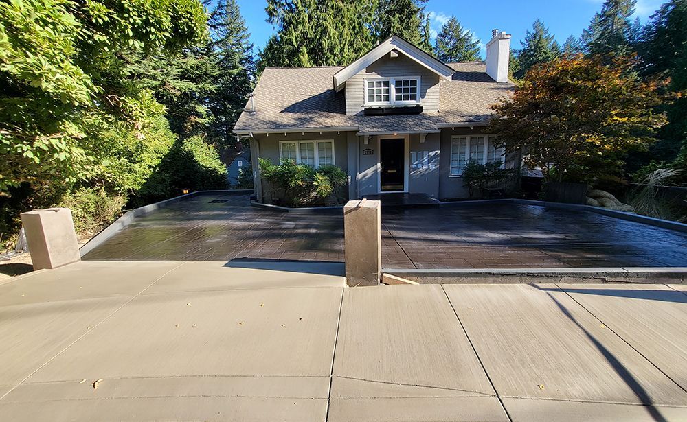 Gray house with a gray driveway, surrounded by trees. Two concrete pillars at the bottom of the driveway.