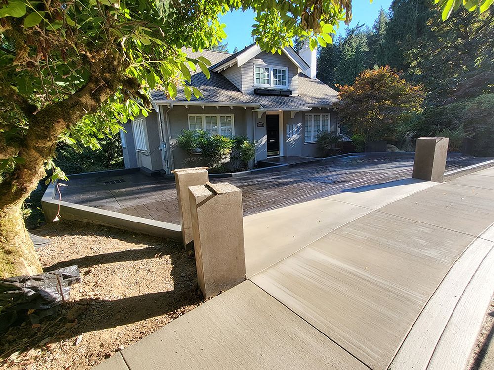 A house with gray siding sits behind a freshly paved concrete driveway. Two concrete pillars stand in front.
