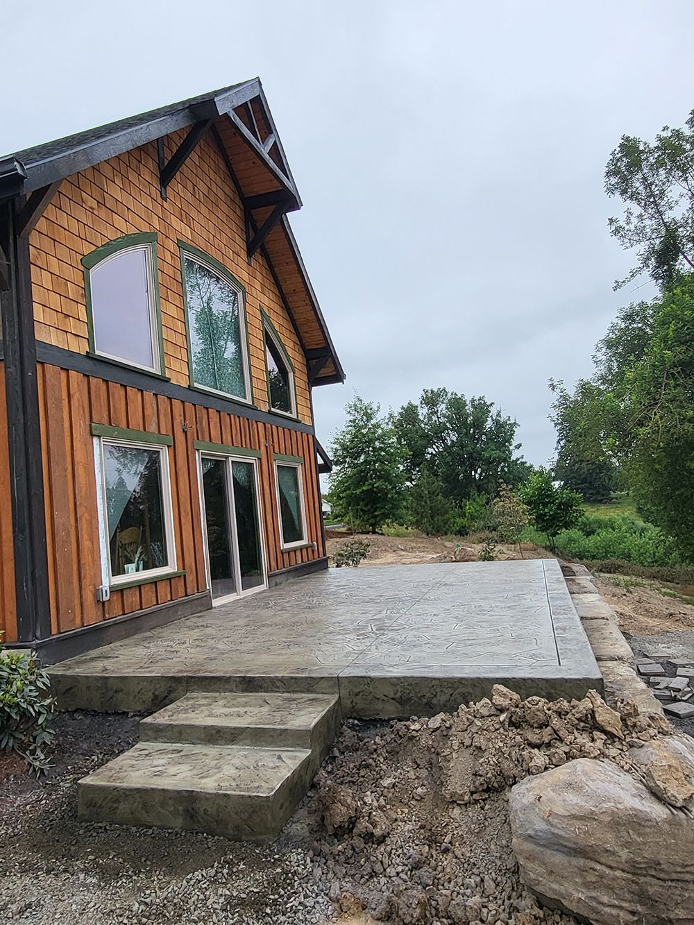 House with a new concrete patio and steps. Brown wood siding, large windows, and surrounding landscaping.