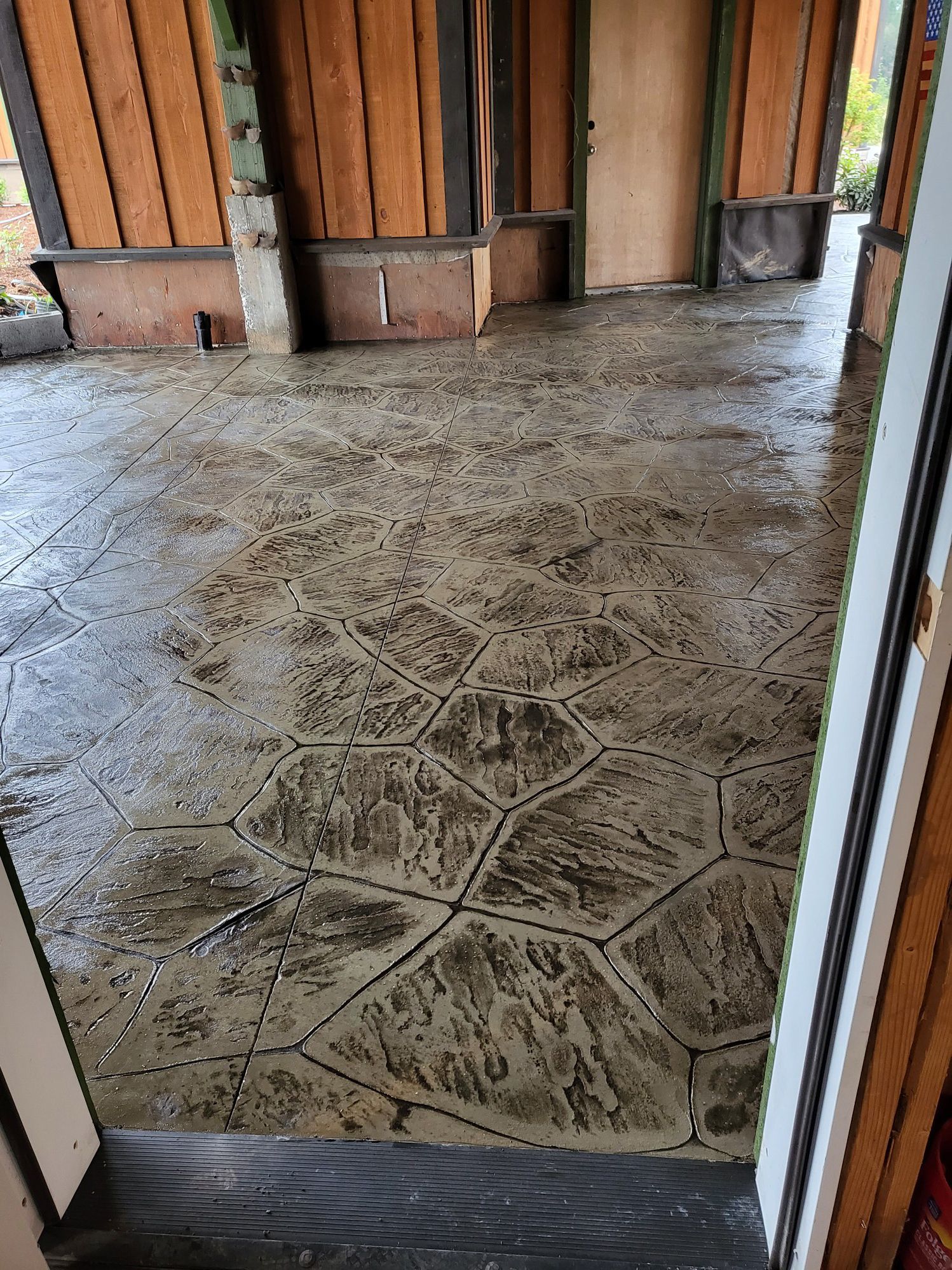 Wet, textured concrete floor with a hexagonal pattern, under a covered outdoor space. Brown wood posts and doorway.
