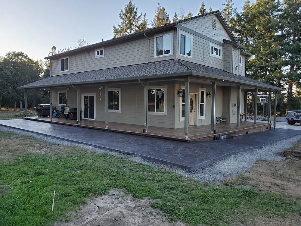 Two-story beige house with wraparound porch and dark gray concrete patio in a grassy setting.