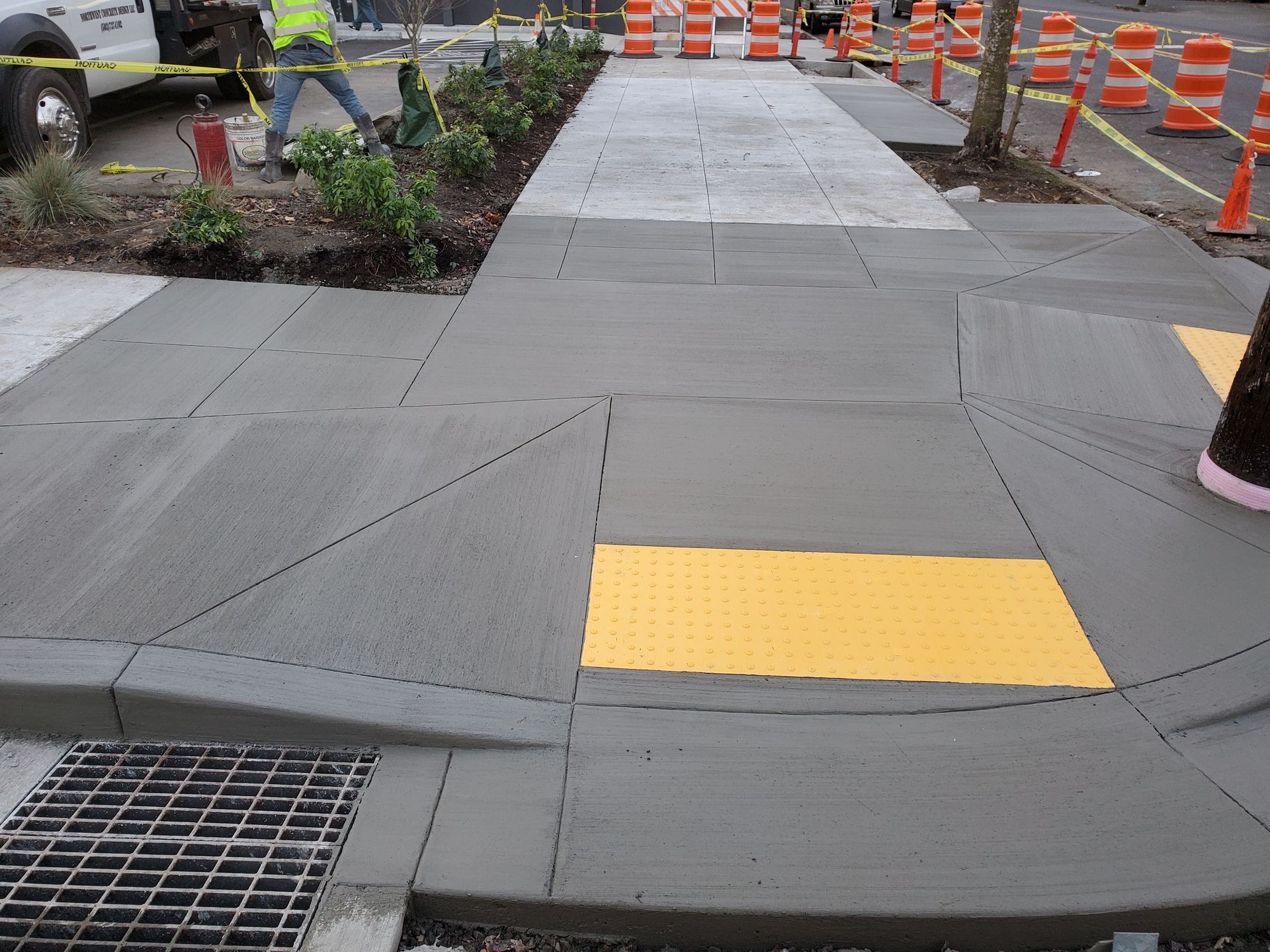 Sidewalk construction: concrete pathway with yellow tactile paving, workers in background, orange cones along street.
