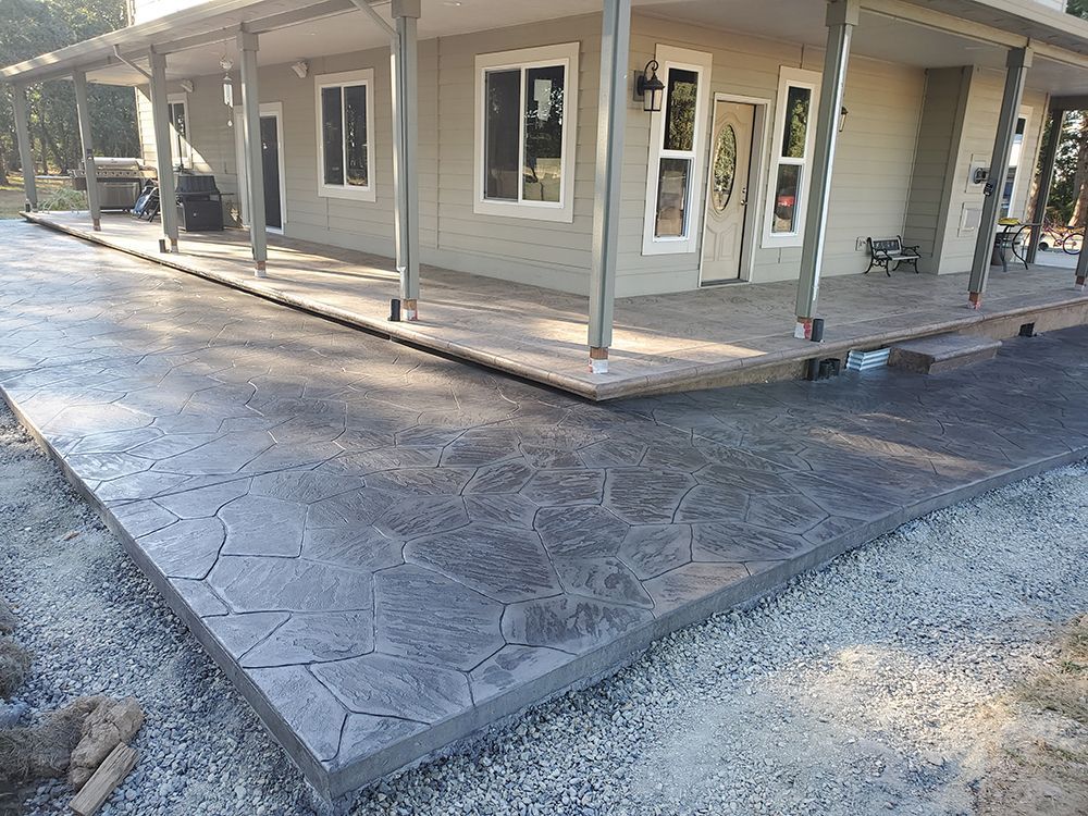 Gray stamped concrete patio in front of a light gray house with a porch and windows.