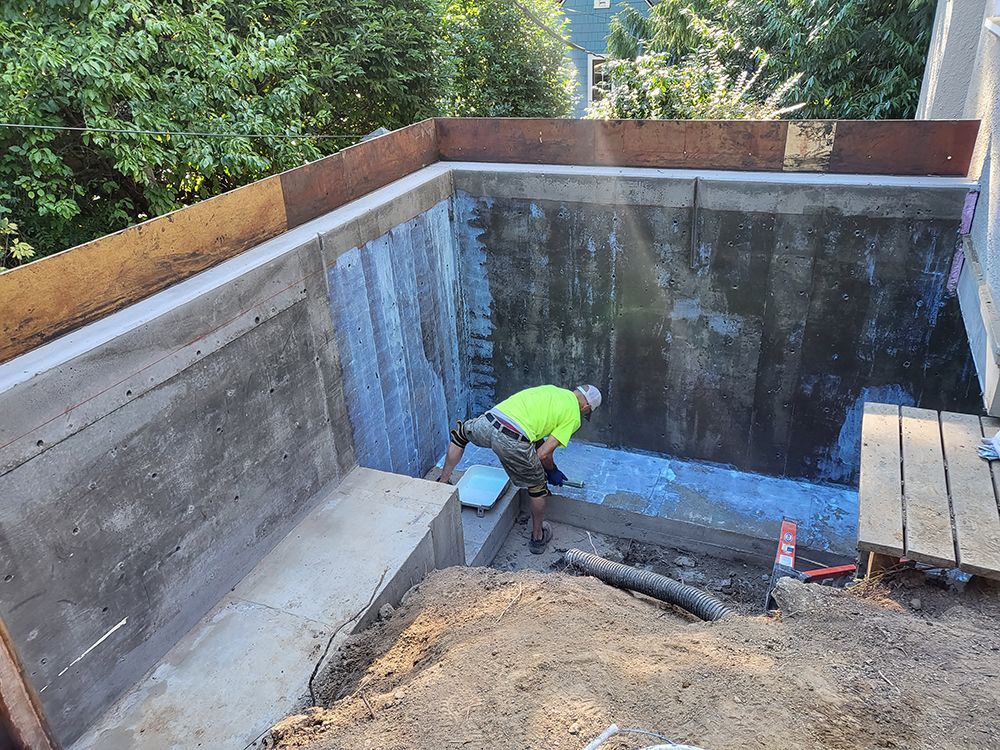 A construction worker in a yellow shirt works inside a concrete pit, possibly waterproofing it.