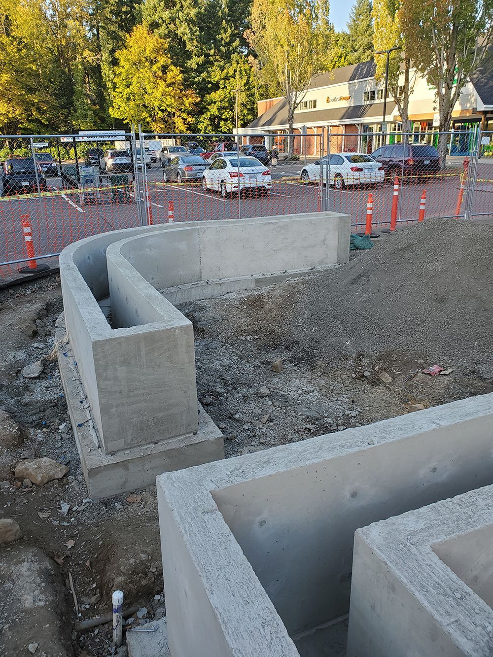 Concrete construction of curved retaining walls with gravel and parked cars in background.