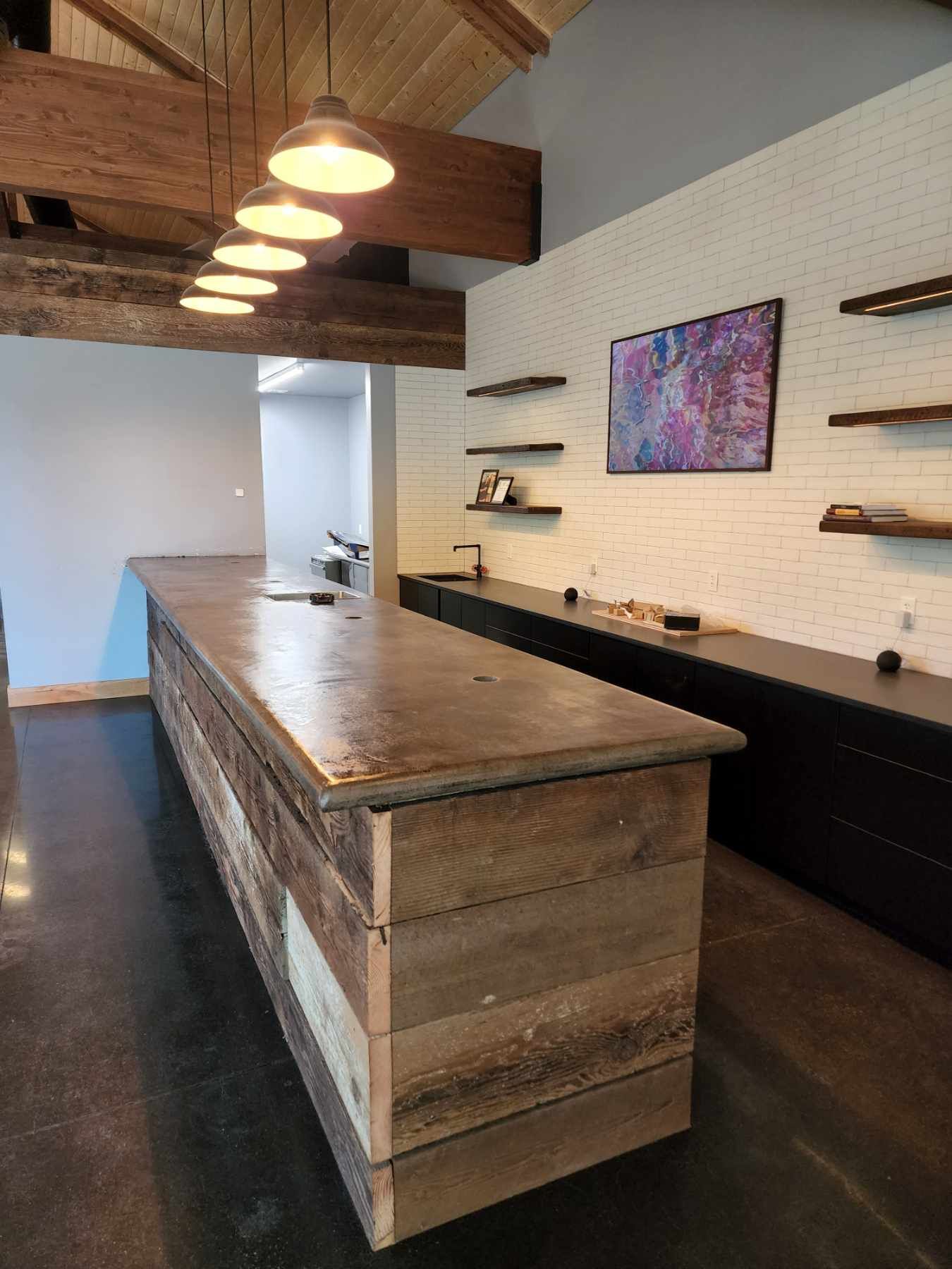 Rustic bar with concrete countertop and reclaimed wood. White brick wall with floating shelves and artwork.