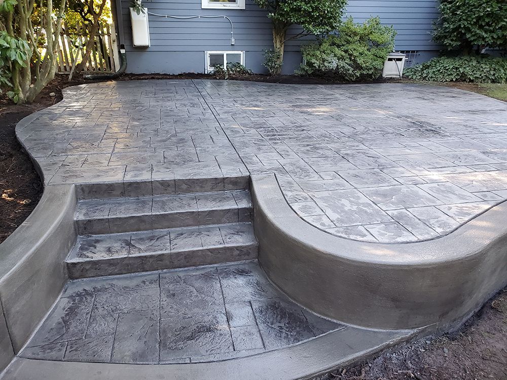 Gray stamped concrete patio with curved edges and steps, next to a blue house with landscaping.