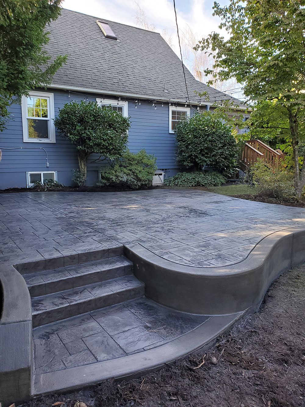 Dark gray stamped concrete patio with curved border and steps leading to a blue house with windows.