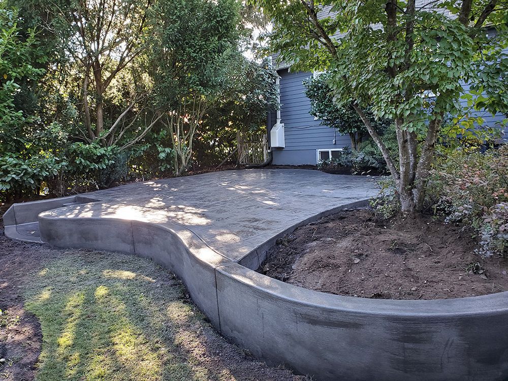 Concrete patio with curved retaining wall, surrounded by trees and greenery.