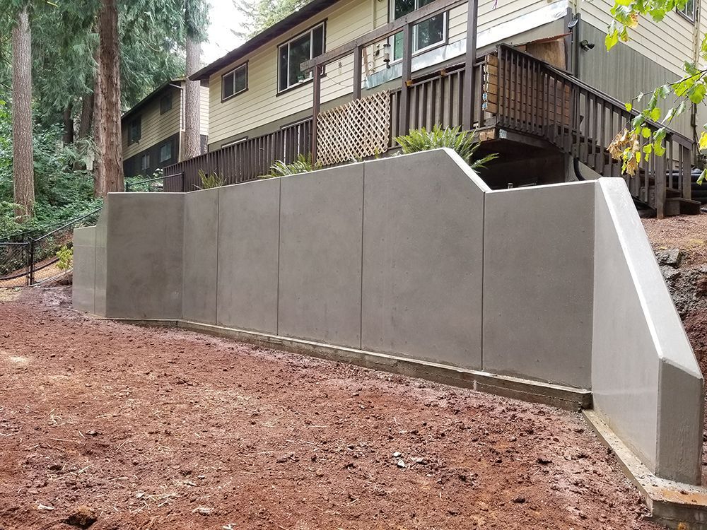 Gray concrete retaining wall in front of a light-colored house with a wooden deck, set on a hillside.