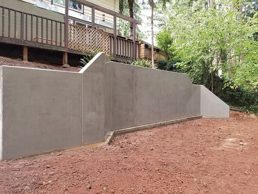 Gray concrete retaining wall in front of a wooden deck, built on a sloped, red-dirt yard.