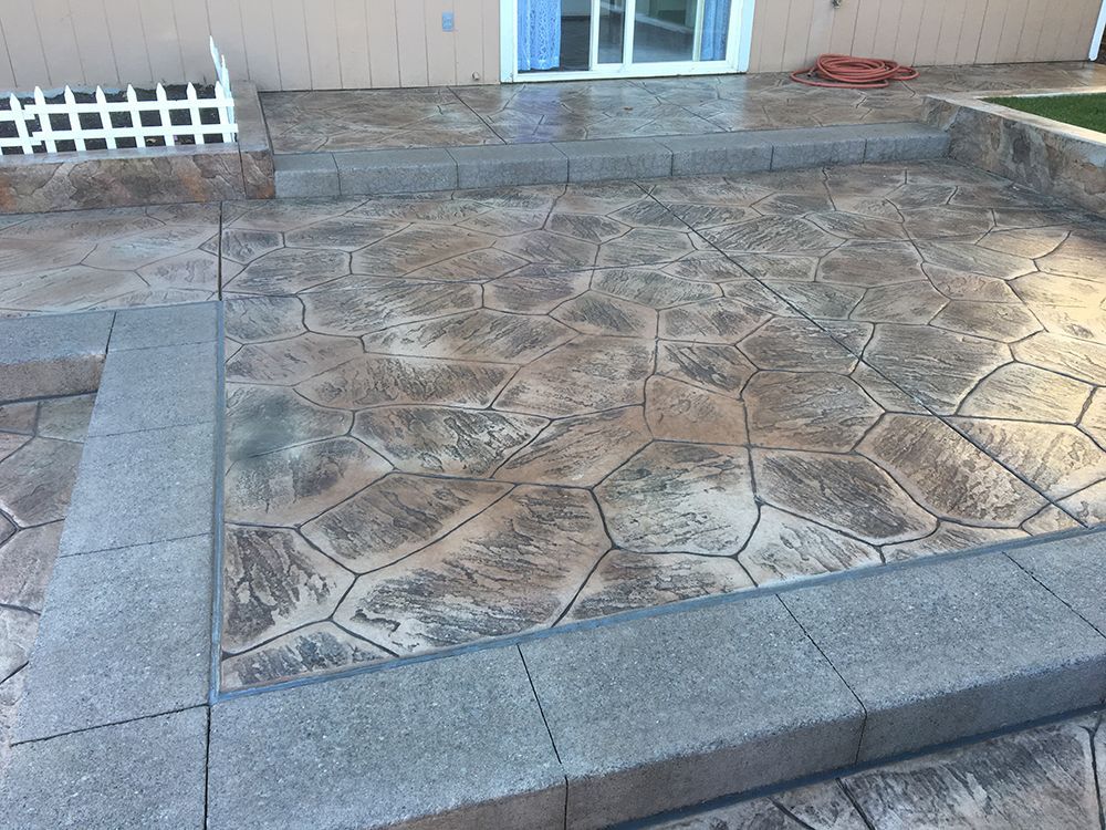 Stamped concrete patio with stone-like pattern, bordered by gray block steps and planters, near a sliding glass door.