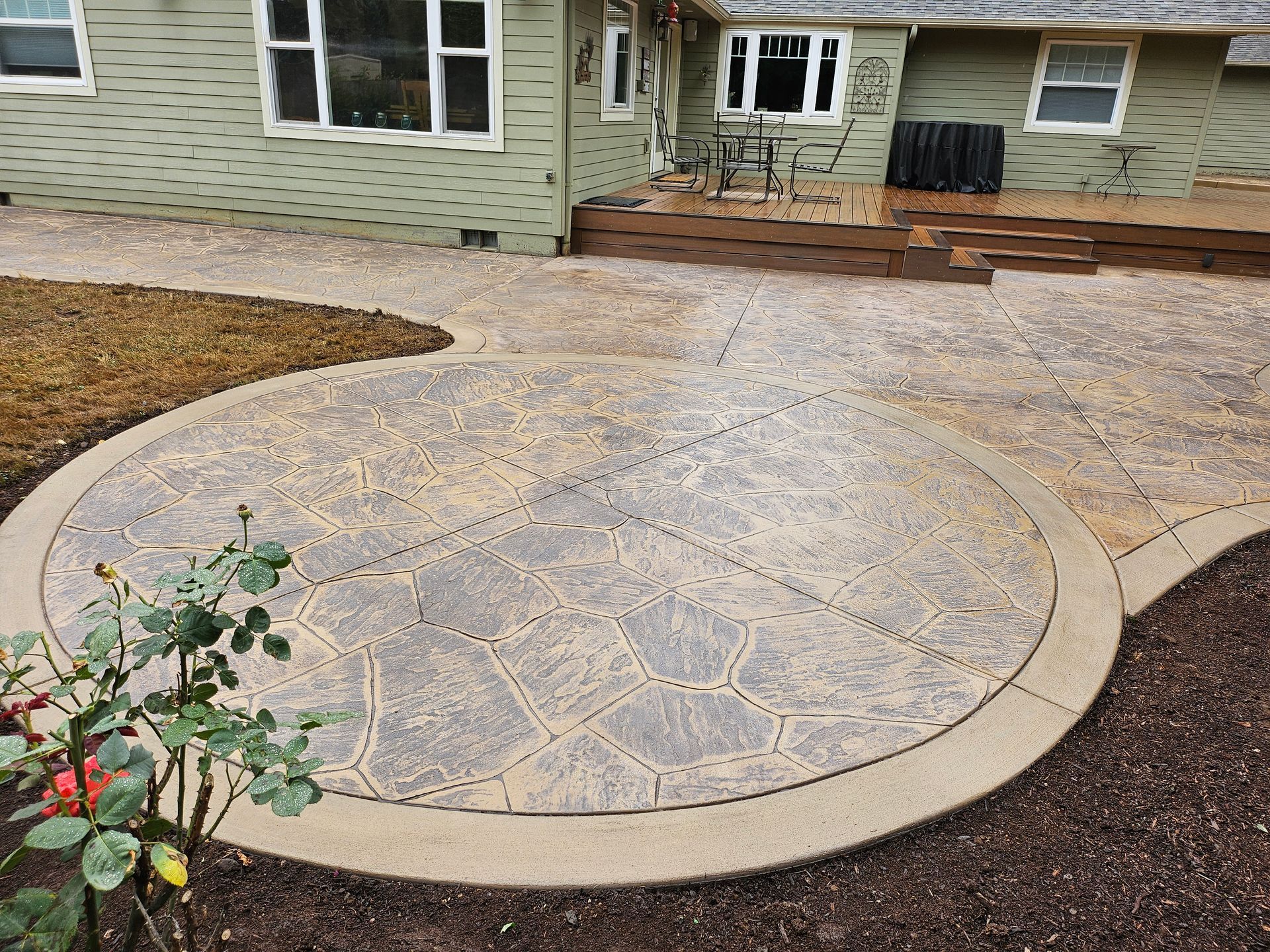 Concrete patio with circular design, brown trim, next to a house with a wooden deck and green siding.
