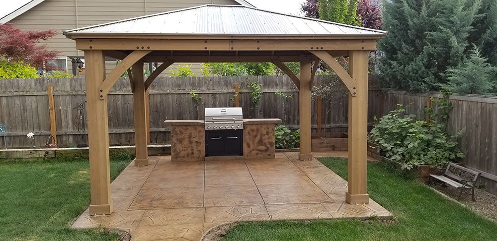 Wooden gazebo with built-in grill in a backyard. Concrete patio and green grass surround it.