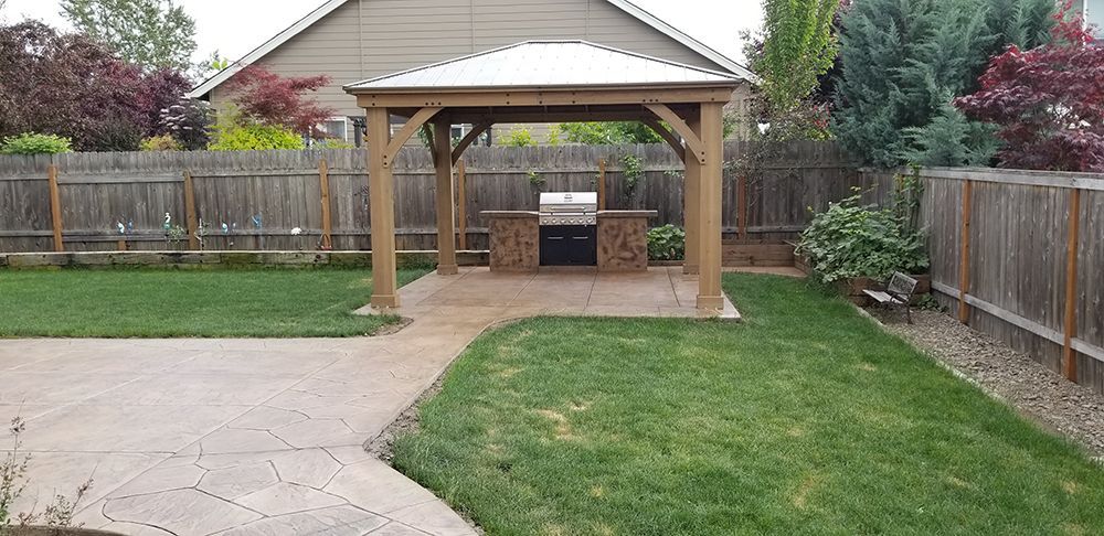 Gazebo with a built-in grill in a backyard with green grass, surrounded by a wooden fence.