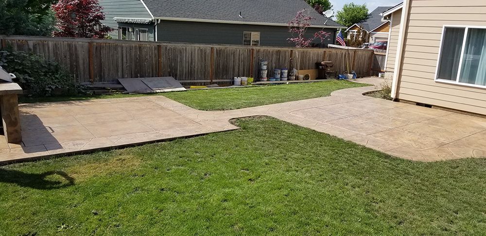 A backyard with a concrete patio, green grass, a wooden fence, and a house.