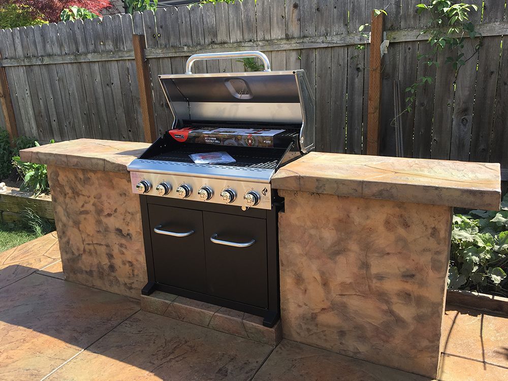 Outdoor grill with open lid on a brown concrete counter, set against a wooden fence.