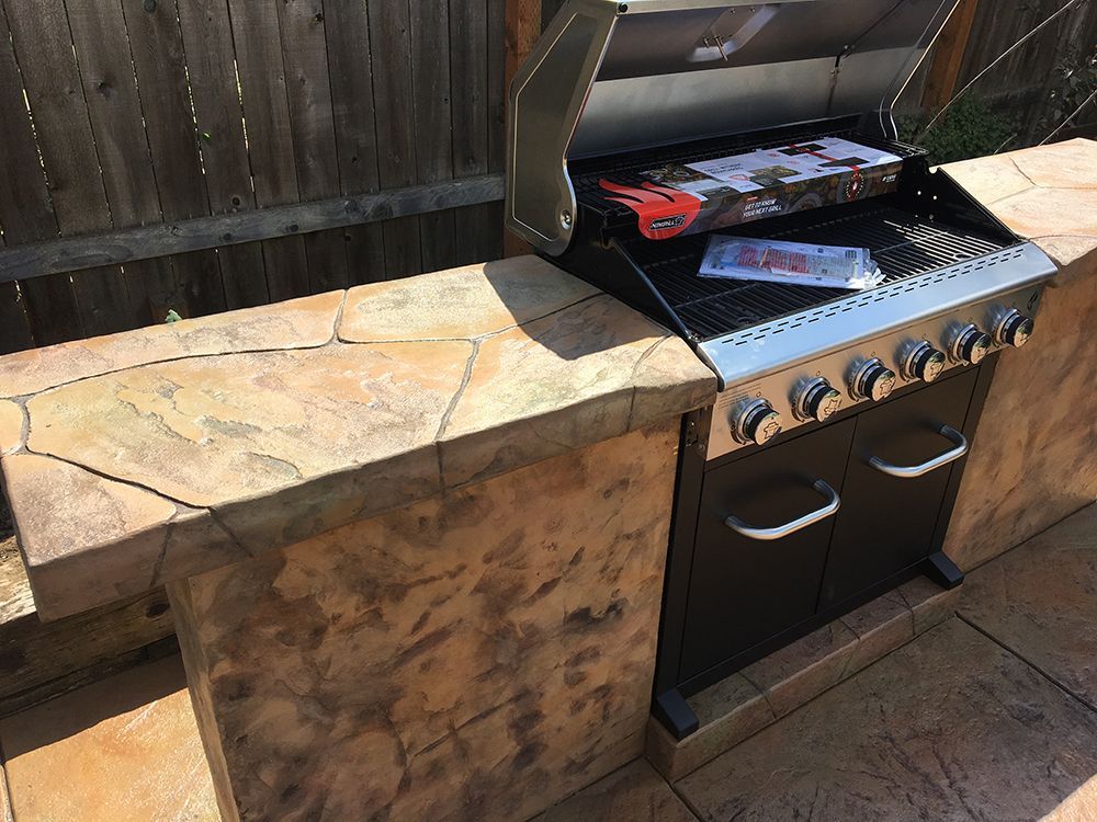 Outdoor built-in grill with brown stone countertop and storage cabinet. A wooden fence is in the background.