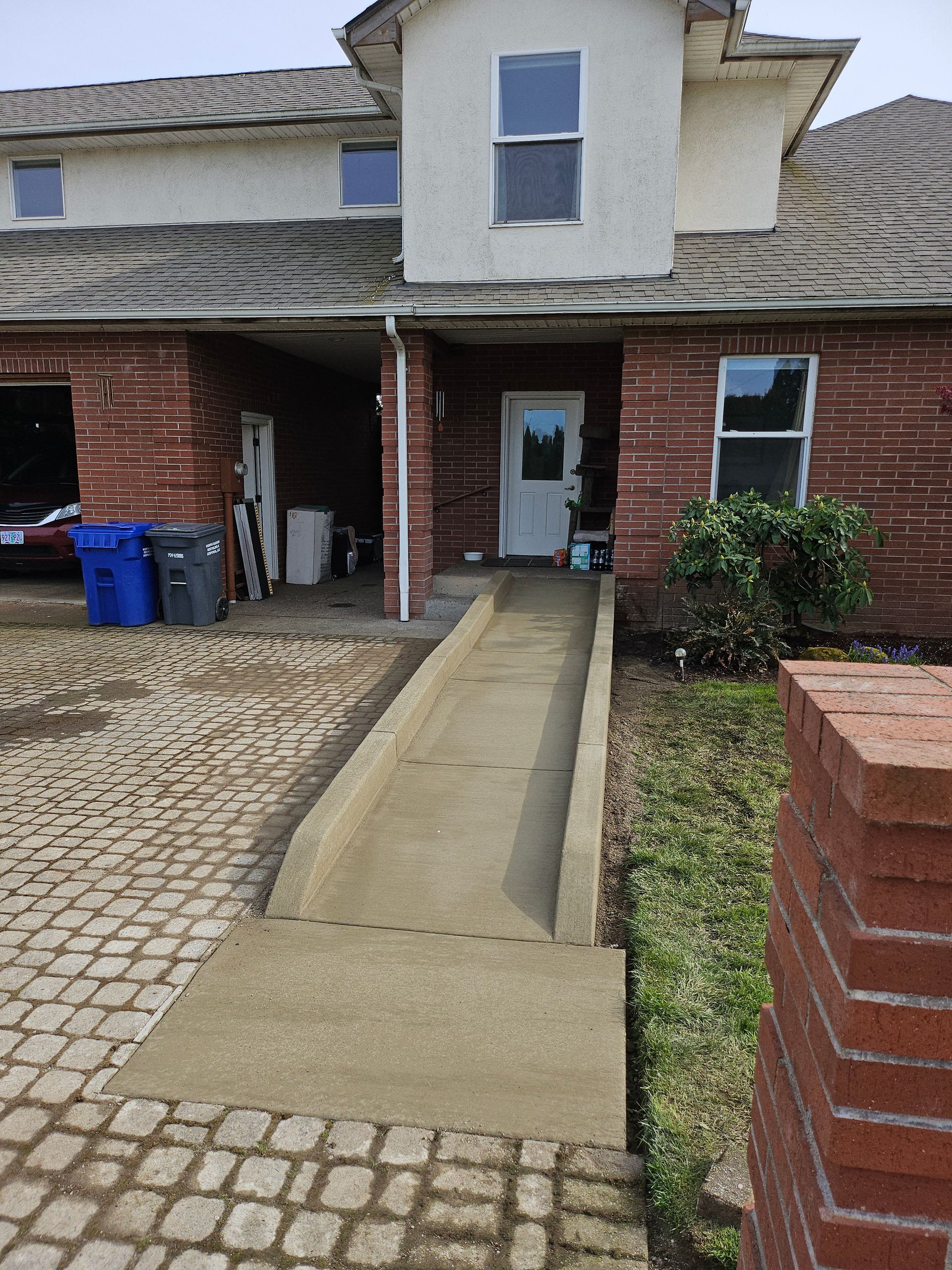 A brick house with a concrete ramp leading to the front door. Landscaping and a brick pillar are visible.