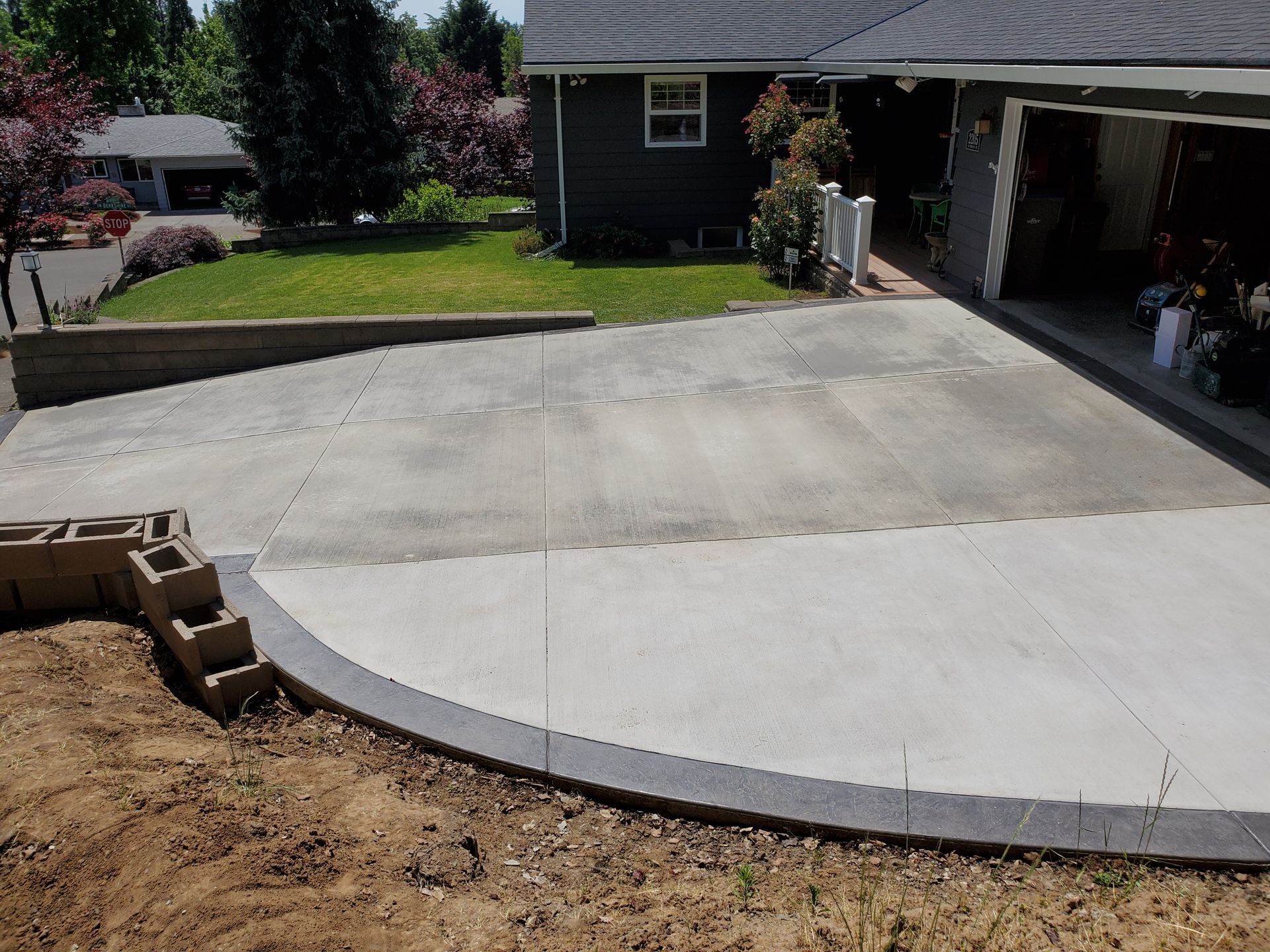 Concrete driveway with gray trim next to a house with a green lawn and shrubs.