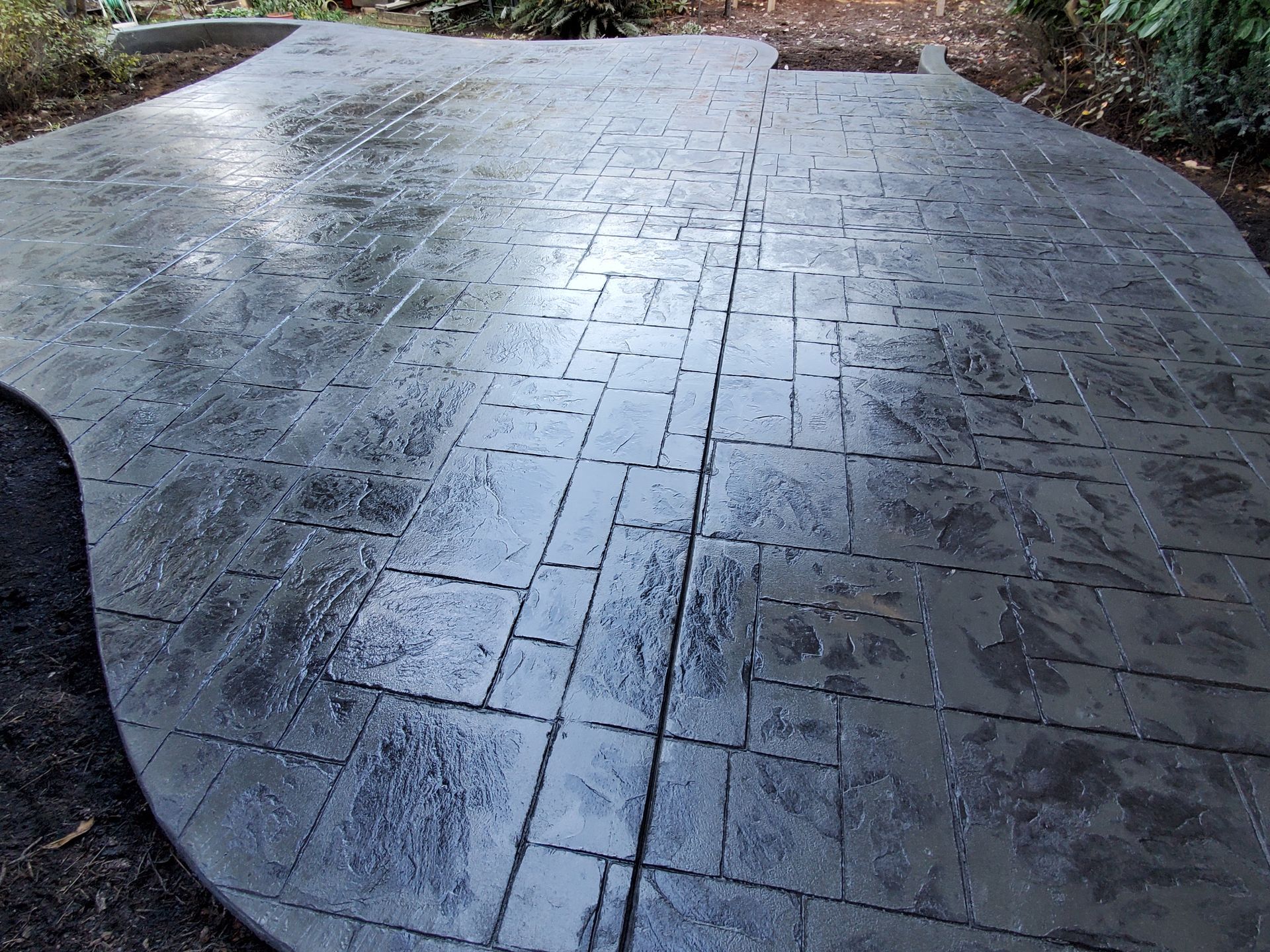 Dark gray stamped concrete patio with textured brick pattern. Wet surface reflects light.
