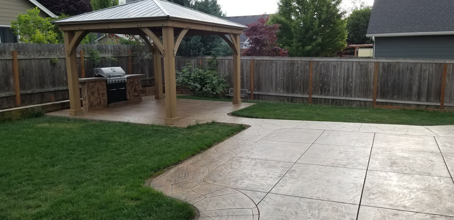 Backyard with a gazebo housing a grill, surrounded by a fence and lawn, and a stamped concrete patio.