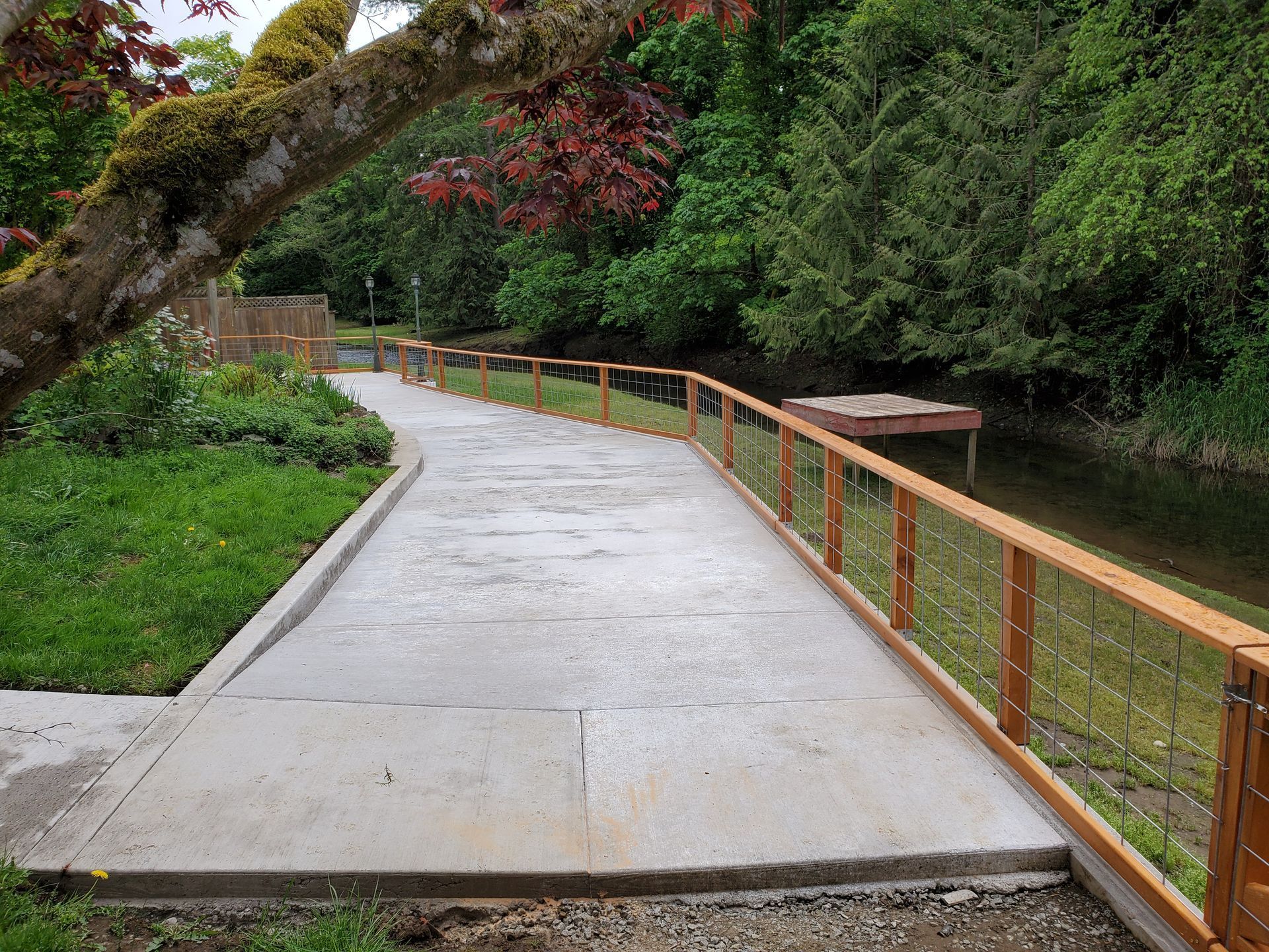 A concrete path with wooden railing alongside a body of water, with trees and greenery in the background.