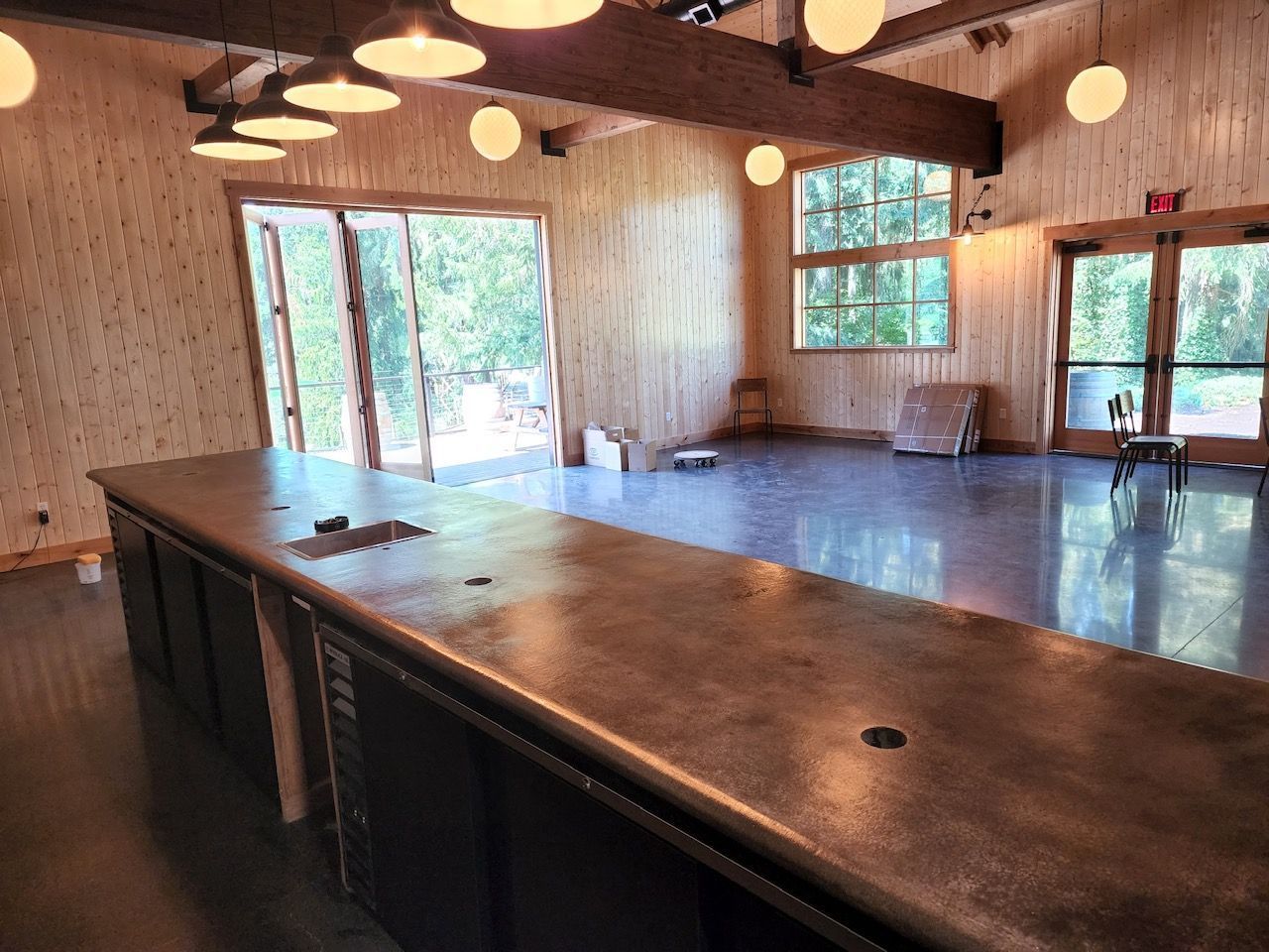 Interior view of a bar with a long counter and windows. Wooden beams and walls, glossy floor, and hanging lights.