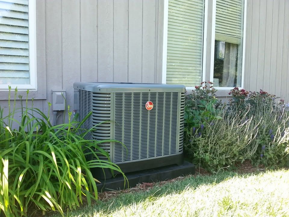 Outdoor air conditioning unit beside a house, surrounded by plants and grass