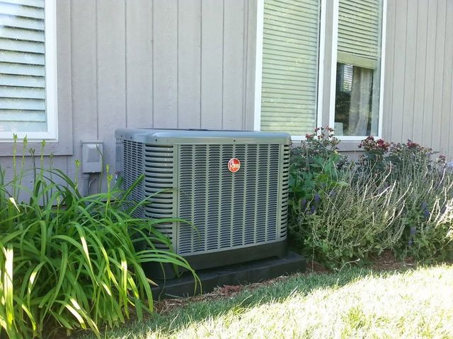 Outdoor air conditioner unit beside a house, surrounded by grass and shrubs.