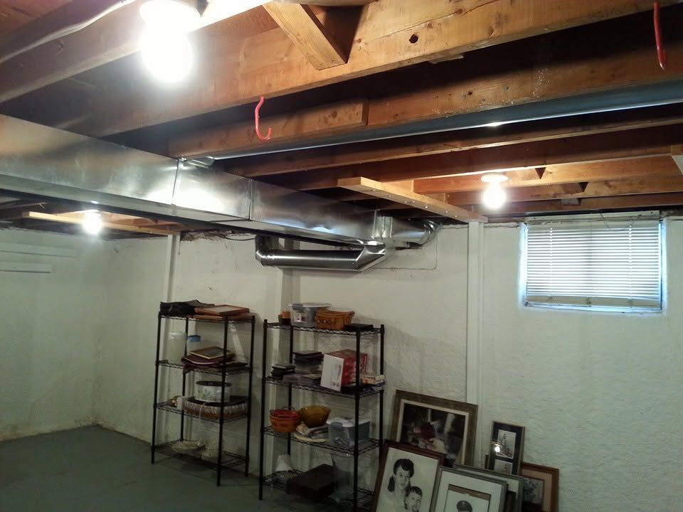 Basement room with exposed beams, storage shelves, and framed photos on the floor by a small window