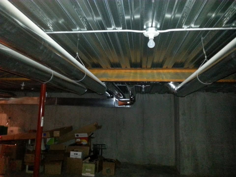 Basement ceiling with exposed silver ducts, pipes, and a single light bulb over stored boxes.