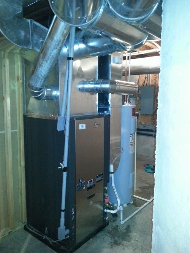 Large HVAC furnace and ductwork in a utility room with metal pipes and insulation.